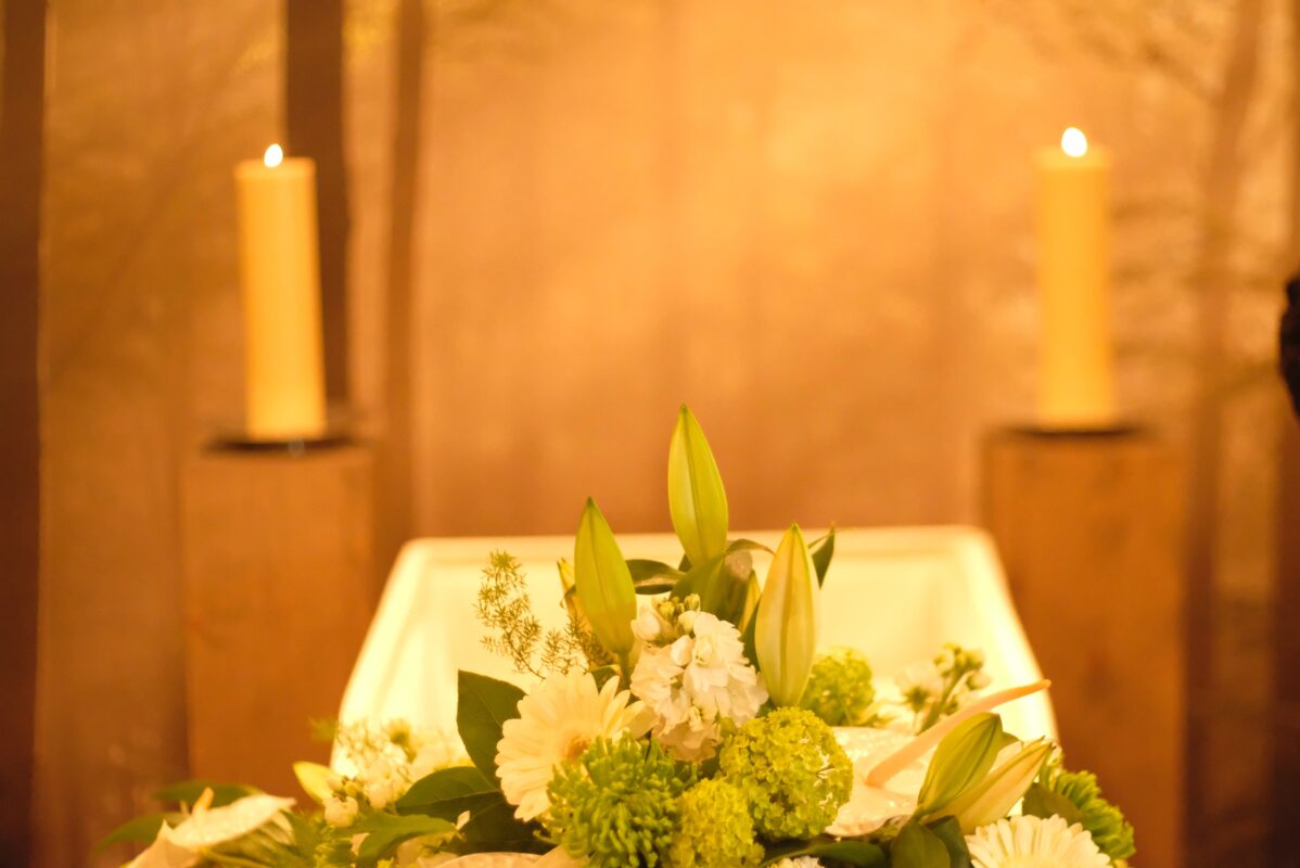 White coffin with white flowers on top, brown background with two long yellow candles.