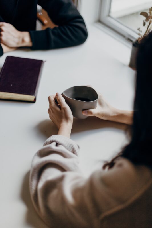 A woman waiting for the embalming process, sitting across a table with a man. The atmosphere looks sad while she drinks coffee and he has a book in Infront of him.